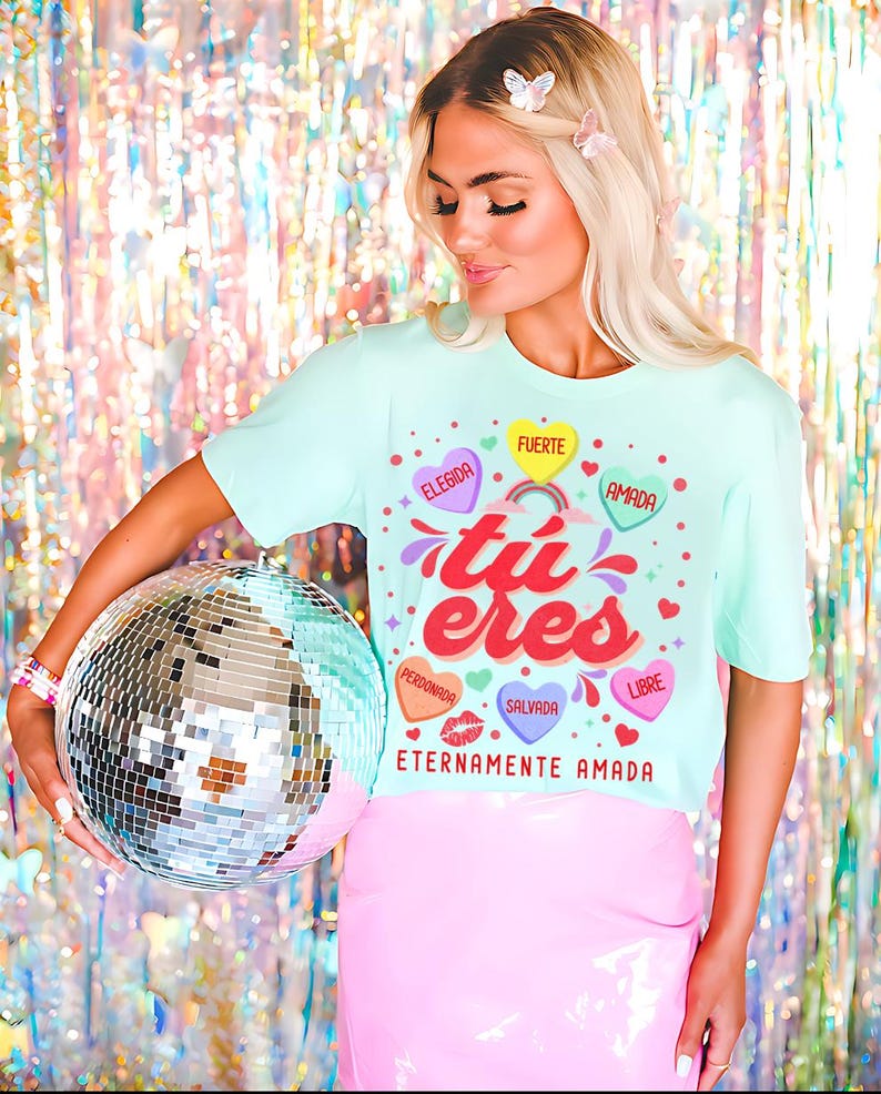 May include: A light blue t-shirt with a graphic design featuring the Spanish words "tu eres" in a colorful font. The graphic also includes hearts with Spanish words like "elegida", "fuerte", "amada", "perdonada", "salvada", and "libre". The shirt also has the text "Eternamente Amada" at the bottom.