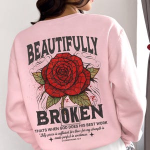 May include: Light pink sweatshirt with a large red rose graphic and the words "BEAUTIFULLY BROKEN". The text "THATS WHEN GOD DOES HIS BEST WORK" and a Bible verse are also printed on the back. The brand logo is at the bottom.