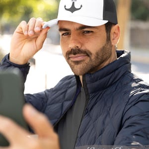 May include: White and black trucker hat with an anchor design and the text "ISAIAH 41:10". The hat has a mesh back and a curved brim. The person in the image is wearing a navy blue quilted jacket.