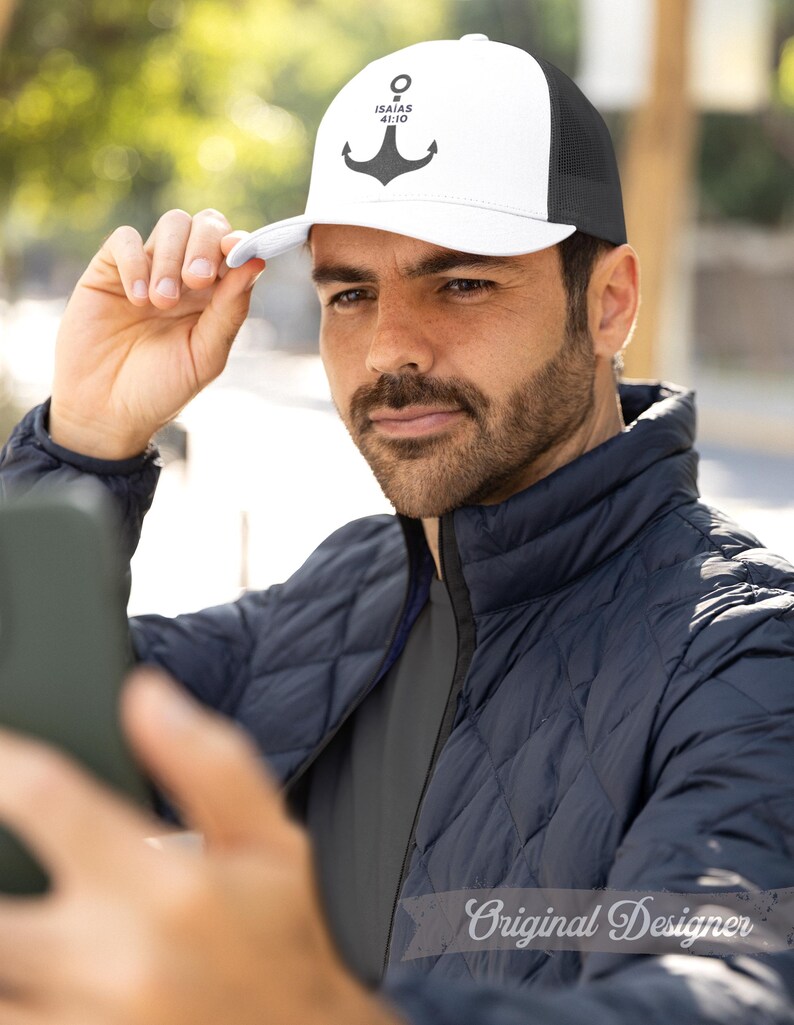 May include: White and black trucker hat with an anchor design and the text "ISAIAH 41:10". The hat has a mesh back and a curved brim. The person in the image is wearing a navy blue quilted jacket.