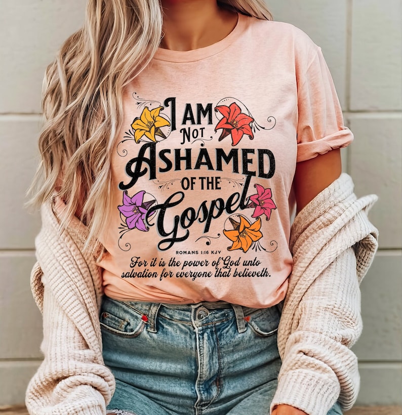 May include: A pink t-shirt with a floral design and the text "I AM NOT ASHAMED OF THE GOSPEL" with the scripture reference "ROMANS 1:16 KJV" and the text "For it is the power of God unto salvation for everyone that believeth."