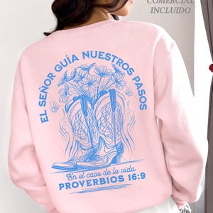 May include: A light pink sweatshirt with a blue graphic design on the back. The design features a pair of cowboy boots, flowers, and text in Spanish. The text includes "EL SE&Ntilde;OR GU&Iacute;A NUESTROS PASOS" and "PROVERBIOS 16:9".