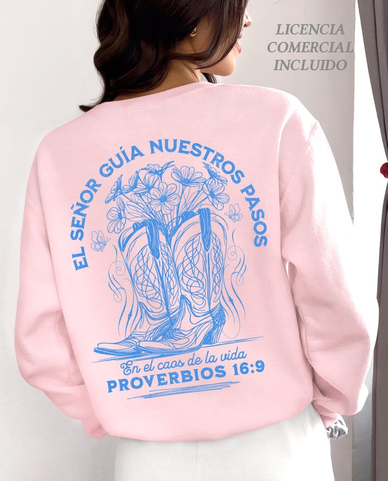 May include: A light pink sweatshirt with a blue graphic design on the back. The design features a pair of cowboy boots, flowers, and text in Spanish. The text includes "EL SE&Ntilde;OR GU&Iacute;A NUESTROS PASOS" and "PROVERBIOS 16:9".
