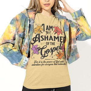 May include: A tan t-shirt with a floral design and the text "I AM NOT ASHAMED OF THE GOSPEL" with the Bible verse "ROMANS 1:16 KJV" and the text "For it is the power of God unto salvation for everyone that believeth."