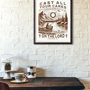 May include: A framed print with the text "CAST ALL YOUR CARES ON THE LORD I PETER 5:7" features a brown and white illustration of a person fishing on a lake with mountains in the background. The print is displayed on a white brick wall.