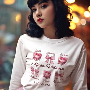 May include: White sweatshirt with a pink bow and the text "Mujer Virtuosa" in a pink script font. The sweatshirt has a design of pink hearts with bows and the words "Noble, Generosa, Cari&ntilde;osa, Trabajadora, Amada, Bendicita" in a pink script font. The text "Proverbios 31" is printed below the design.