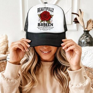 May include: A black and white trucker hat with the words "BEAUTIFULLY BROKEN" above a red rose graphic. The hat also features the text "Thats when God does his best work". The hat has a mesh back.