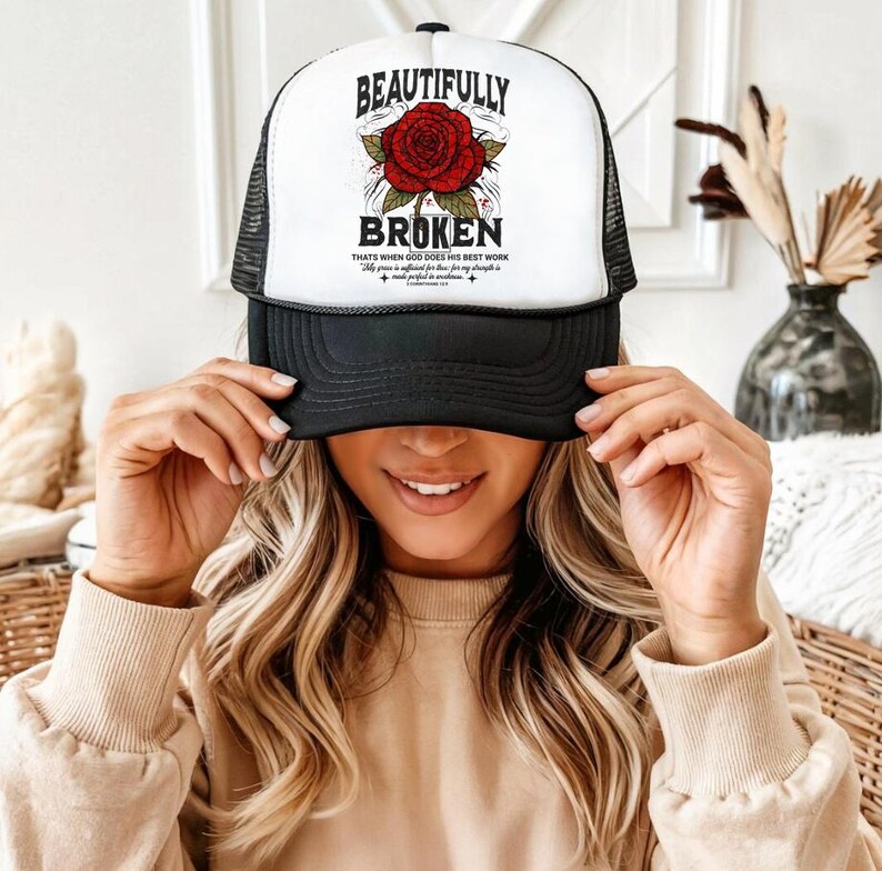 May include: A black and white trucker hat with the words "BEAUTIFULLY BROKEN" above a red rose graphic. The hat also features the text "Thats when God does his best work". The hat has a mesh back.