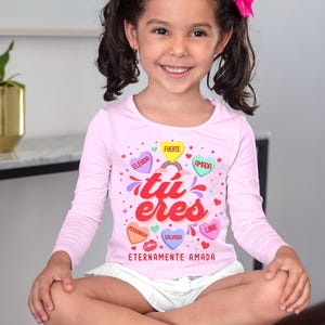 May include: A pink long-sleeved shirt with a graphic design of hearts with Spanish words on them. The words are "Elegida", "Fuerte", "Amada", "Perd&oacute;nan", "Salva", and "Libre". The shirt also has the words "tu eres" and "Eternamente Amada" printed on it.