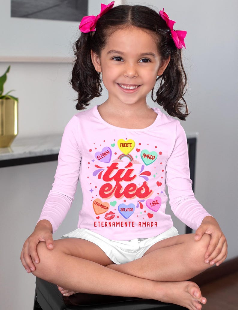 May include: A pink long-sleeved shirt with a graphic design of hearts with Spanish words on them. The words are "Elegida", "Fuerte", "Amada", "Perd&oacute;nan", "Salva", and "Libre". The shirt also has the words "tu eres" and "Eternamente Amada" printed on it.