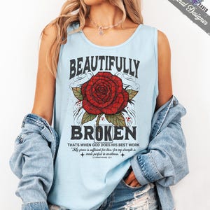 May include: Light blue tank top with the words "BEAUTIFULLY BROKEN" in black, featuring a red rose graphic. The text "THATS WHEN GOD DOES HIS BEST WORK" and a Bible verse are also printed on the shirt.