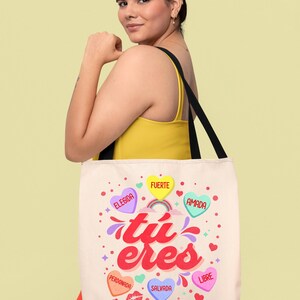 May include: A white tote bag with a pink, yellow, and blue design. The bag has the words "tu eres" in a pink and red font. The bag also has several hearts with words on them, including "elegida", "fuerte", "amada", "perdonada", "salvada", and "libre". The bag also has the words "eternamente amada" at the bottom.