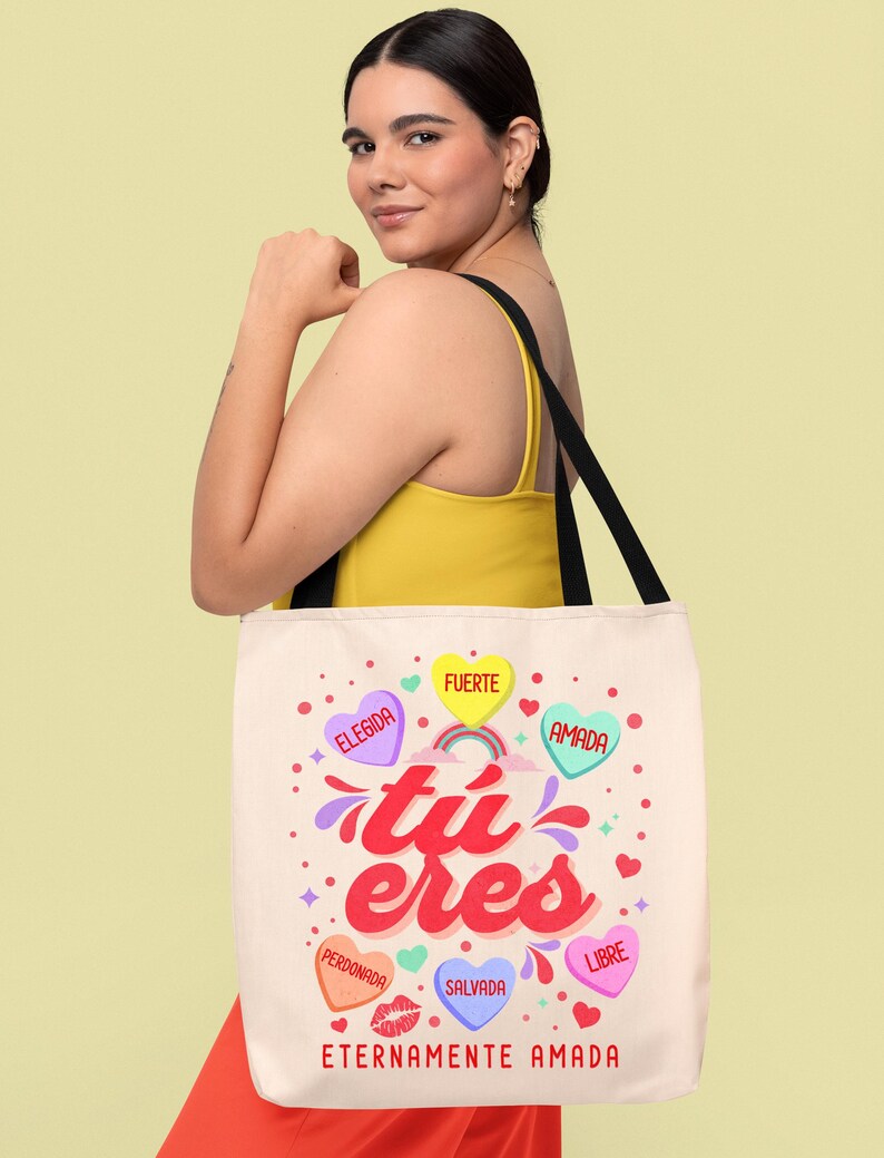 May include: A white tote bag with a pink, yellow, and blue design. The bag has the words "tu eres" in a pink and red font. The bag also has several hearts with words on them, including "elegida", "fuerte", "amada", "perdonada", "salvada", and "libre". The bag also has the words "eternamente amada" at the bottom.