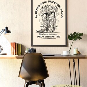 May include: A framed black and white print featuring cowboy boots filled with flowers. The text reads "EL SE&Ntilde;OR GU&Iacute;A NUESTROS PASOS" and "En el caos de la vida PROVERBIOS 16:9". The print is displayed on a wall above a desk with a chair.