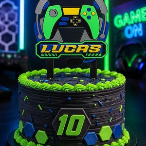 Puede incluir: Un pastel redondo con glaseado negro, bordes verdes y chispas azules y verdes. Un adorno muestra un mando de juego verde, auriculares y el nombre LUCAS en amarillo. Un hexágono negro con el número 10 en verde se encuentra en la parte frontal.