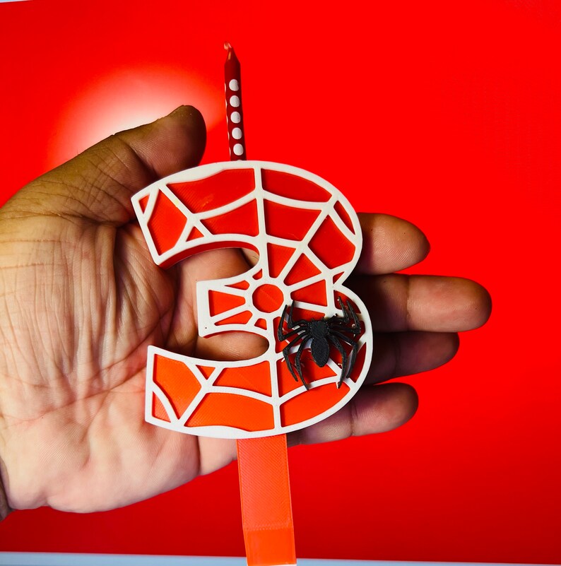 May include: A red and white spiderweb-shaped candle with a black spider on it. The candle is shaped like the number three.