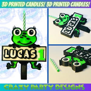 Puede incluir: Un adorno para pastel de cumpleaños de rana verde impreso en 3D con un letrero que dice "Lucas 1". La rana sostiene el letrero con sus manos. El adorno para pastel está diseñado para un pastel de cumpleaños.