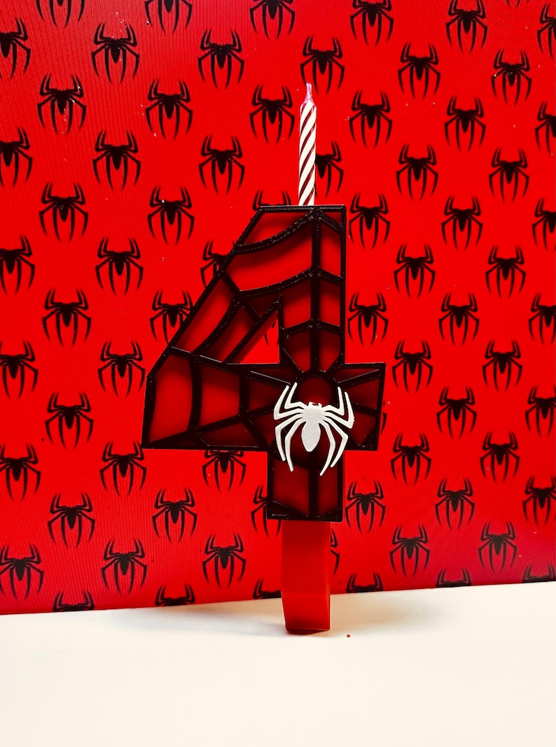 May include: A red and black number four candle with a spider web design and a white spider in the centre. The candle is on a red background with a pattern of black spiders.