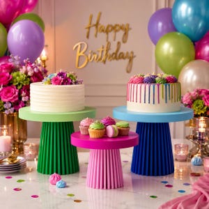 May include: A festive birthday scene with three tiered cake stands in green, pink, and blue, each holding a cake or cupcakes. The cakes are decorated with flowers and colorful frosting. Balloons and the words "Happy Birthday" complete the celebratory setting.