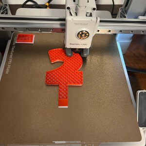 May include: A red 3D printed object with a grid pattern is being printed on a textured PEI plate on a 3D printer. The printer is labelled "Bambu Lab".