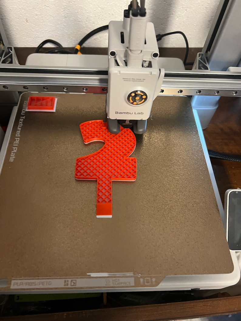 May include: A red 3D printed object with a grid pattern is being printed on a textured PEI plate on a 3D printer. The printer is labelled "Bambu Lab".