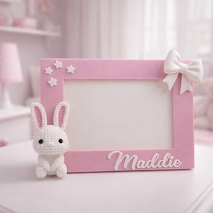 Puede incluir: Un marco de fotos rosa con el nombre "Maddie" en escritura blanca, decorado con estrellas blancas y un lazo. Un pequeño conejo blanco tejido se sienta junto al marco. El marco está sobre una superficie blanca.