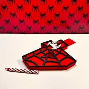 May include: A red and black 3D printed number one with a white spider on top. The number one is shaped like a spider web. A red and white striped candle is in the foreground.