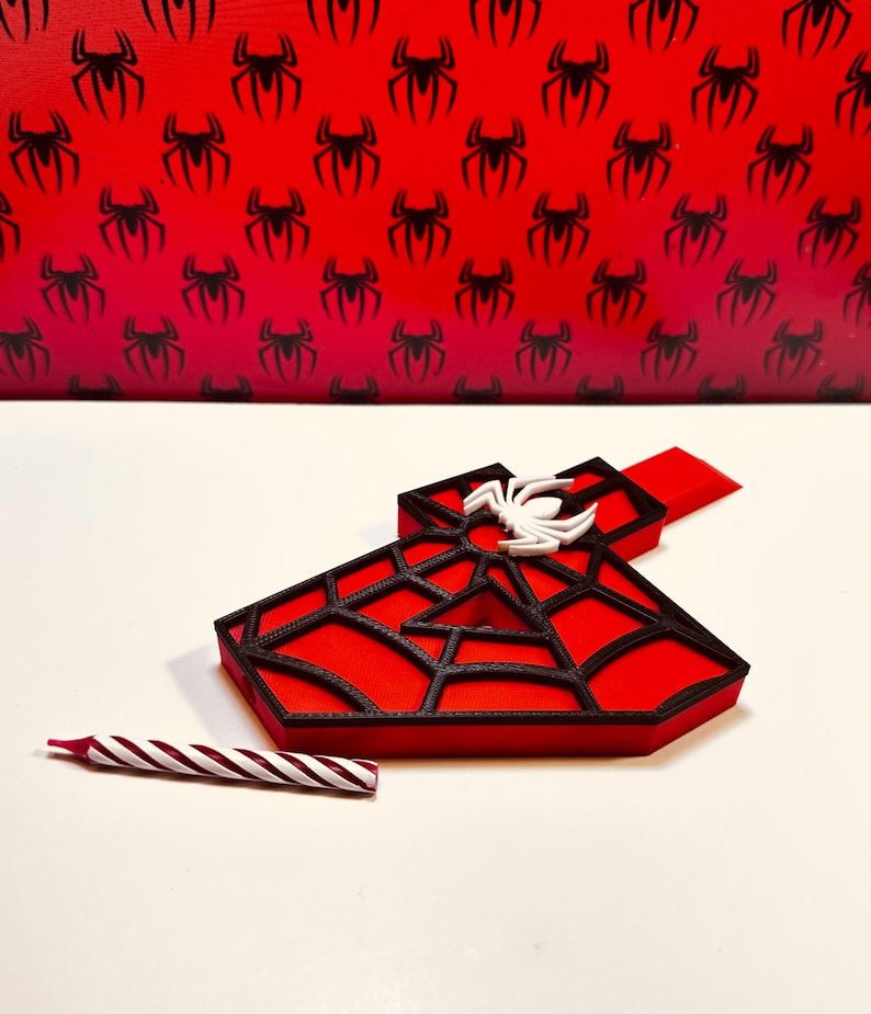 May include: A red and black 3D printed number one with a white spider on top. The number one is shaped like a spider web. A red and white striped candle is in the foreground.