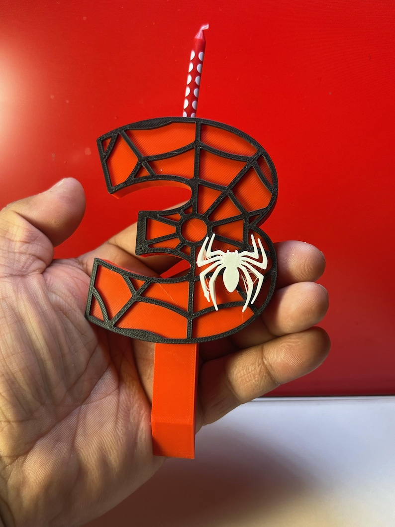 May include: A red and black 3D printed number three candle with a spider web design and a white spider on the front. The candle is on a red background.