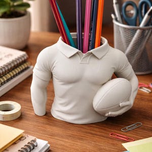 May include: A white rugby-themed pencil holder in the shape of a torso, holding a football. The holder contains several coloured pencils. The desk also has a roll of tape, notebooks, and paperclips.