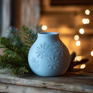 Pode incluir: Um vaso de cerâmica azul claro com desenhos de flocos de neve em relevo. O vaso é redondo com uma base larga e um pescoço estreito. Um ramo de sempre-viva está à esquerda do vaso. O fundo tem um efeito bokeh desfocado.