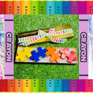 Puede incluir: Cuatro crayones en forma de rompecabezas de color amarillo, azul, naranja y rojo, dispuestos en una fila en una caja dorada. La caja está atada con una cinta blanca con un patrón colorido. La caja está sentada sobre una superficie verde cubierta de hierba.
