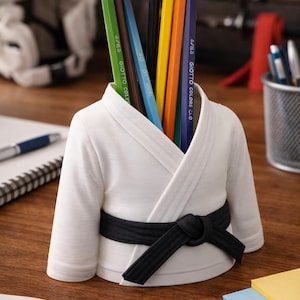 May include: A white pencil holder in the shape of a karate uniform, featuring a black belt. It's filled with various coloured pencils: green, brown, blue, black, yellow, orange, and purple. The pencils are labelled with the brand "GIOTTO COLORS C.O."
