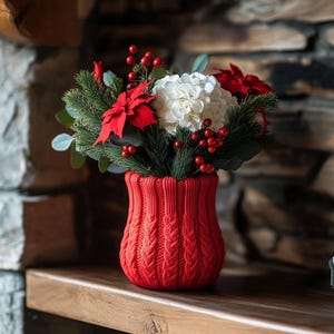 Op de afbeelding: Een feestelijk bloemstuk in een rode gebreide vaas. Het boeket bevat witte hortensia's, rode poinsettia's, groene dennentakken en rode bessen. De vaas heeft een textuur met kabelbreisel, wat een gezellige, seizoensgebonden touch toevoegt.