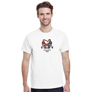 Könnte beinhalten: Weißes T-Shirt mit einer Grafik von zwei Tukanen, die Schach spielen, darunter der Text "ToucanBru 1998". Die Tukane sind in Rot und Blau gekleidet und sitzen an einem Tisch mit Getränken. Das T-Shirt wird von einer Person getragen.