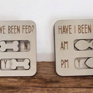 May include: Two wooden pet feeding indicators. One reads "HAVE WE BEEN FED?" with bone-shaped sliders for AM and PM. The other reads "HAVE I BEEN FED?" with fish-shaped sliders. Both are on a wooden surface.