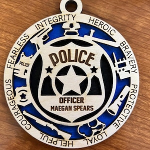 May include: A wooden police badge ornament with a blue and black background. The ornament features the word "POLICE" and the name "OFFICER MAEGAN SPEARS". The design includes stars, guns, and words like "Courageous" and "Heroic".