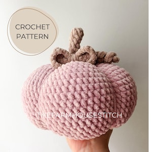 May include: A crocheted pumpkin with a stem made of the same yarn. The pumpkin is a soft pink color and has a textured surface. The text "THE FARMHOUSESTITCH" is visible on the pumpkin.