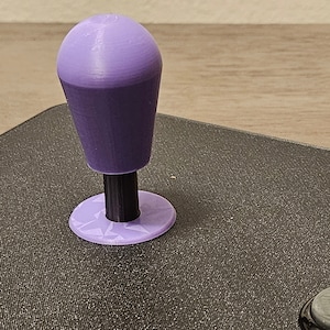 May include: Purple arcade joystick with a round top and a base with a starburst pattern.