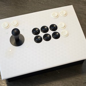 May include: White arcade game controller with a black base. It features a black joystick and several white and black buttons. The buttons are arranged in a classic arcade layout.