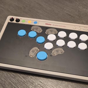 Peut inclure: Un contrôleur d'arcade rectangulaire avec un cadre gris et un panneau supérieur noir. Il comporte des boutons bleus et blancs, des boutons directionnels transparents et des boutons de contrôle. Le texte "8BitDo Arcade Controller" est imprimé en haut.