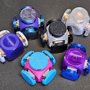 May include: Six colorful, 3D printed, geometric toys with spinning parts. Each toy has a different color and design. The toys are made of plastic and have a smooth, glossy finish.