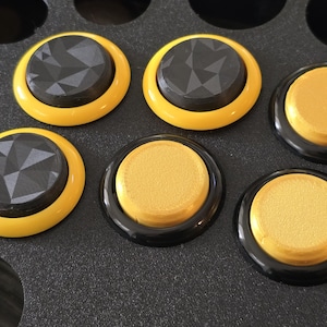May include: Six round buttons with black and yellow accents. Three buttons have a black centre with a textured diamond pattern and a yellow outer ring. Three buttons have a gold centre and a black outer ring.