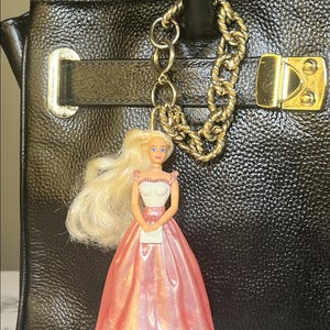 May include: A black leather handbag with a gold chain and clasp, adorned with a doll charm. The doll has blonde hair and wears a pink and white dress, holding a small white purse. The charm is attached to the bag with a gold chain.