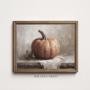 May include: A framed oil painting print featuring a single orange pumpkin resting on a white cloth surface with a small cluster of dried flowers. The artwork is enclosed in a thin, metallic-toned frame and includes the text SOLASTA PRINT below.