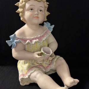 May include: A porcelain figurine of a young girl with blonde hair, wearing a yellow and pink dress with a ruffled trim and blue bows. She is sitting with her legs crossed and holding a small cup.
