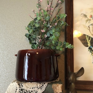 May include: A dark brown ceramic planter with a rounded shape and two handles. The planter is filled with a leafy green plant with small pink flowers. The planter is sitting on top of a stack of old books and a crocheted doily. A small bronze mouse figurine is sitting on the table next to the planter.