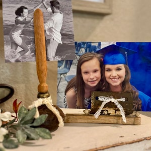 May include: Two framed photos on a wooden table. The photo on the left is a black and white photo of a couple dancing in the water. The photo on the right is a color photo of two girls, one wearing a blue graduation cap. The photos are held up by wooden dowels with a decorative base.