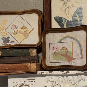 May include: Two framed cross-stitch embroidery pieces with a rustic farmhouse theme. The first piece depicts a small house with a chimney, a tree, and a squirrel. The second piece shows a house with a rainbow above it and a path leading to the house.