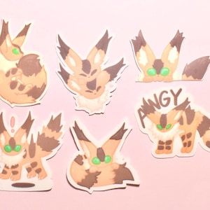 May include: A set of six die-cut stickers featuring a tan and brown creature with large pointed ears and bright green eyes in various poses, including one sticker with the text ANGY written in a dark brown font above the creature.